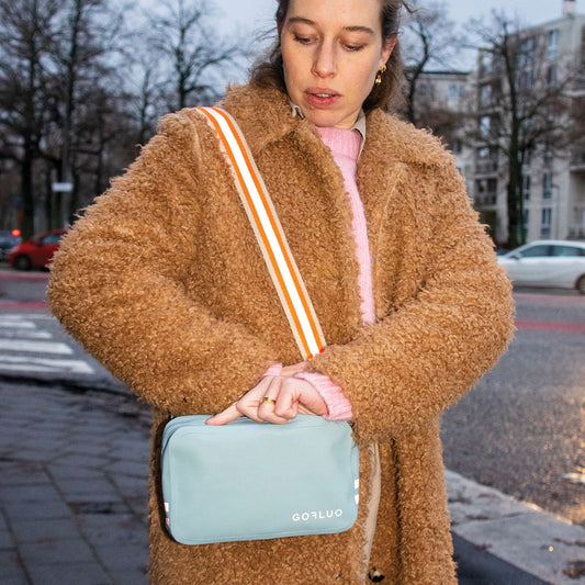 Woman in a brown coat holding a light blue bag with a blurred street background