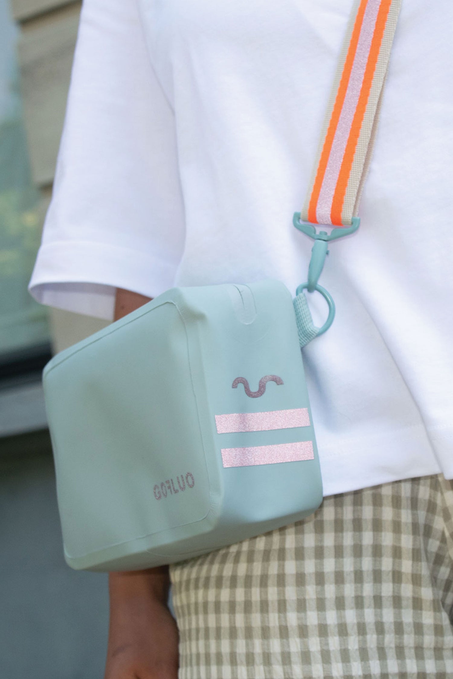 Light blue crossbody bag with pink accents held by a person wearing a white shirt and plaid shorts.