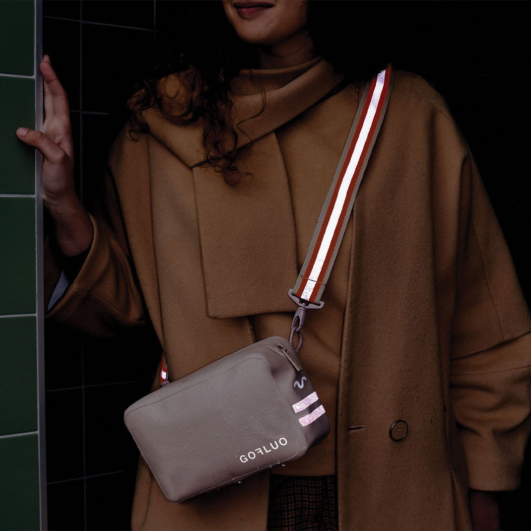 Person wearing a brown coat with a reflective bag strap, standing against a dark background.