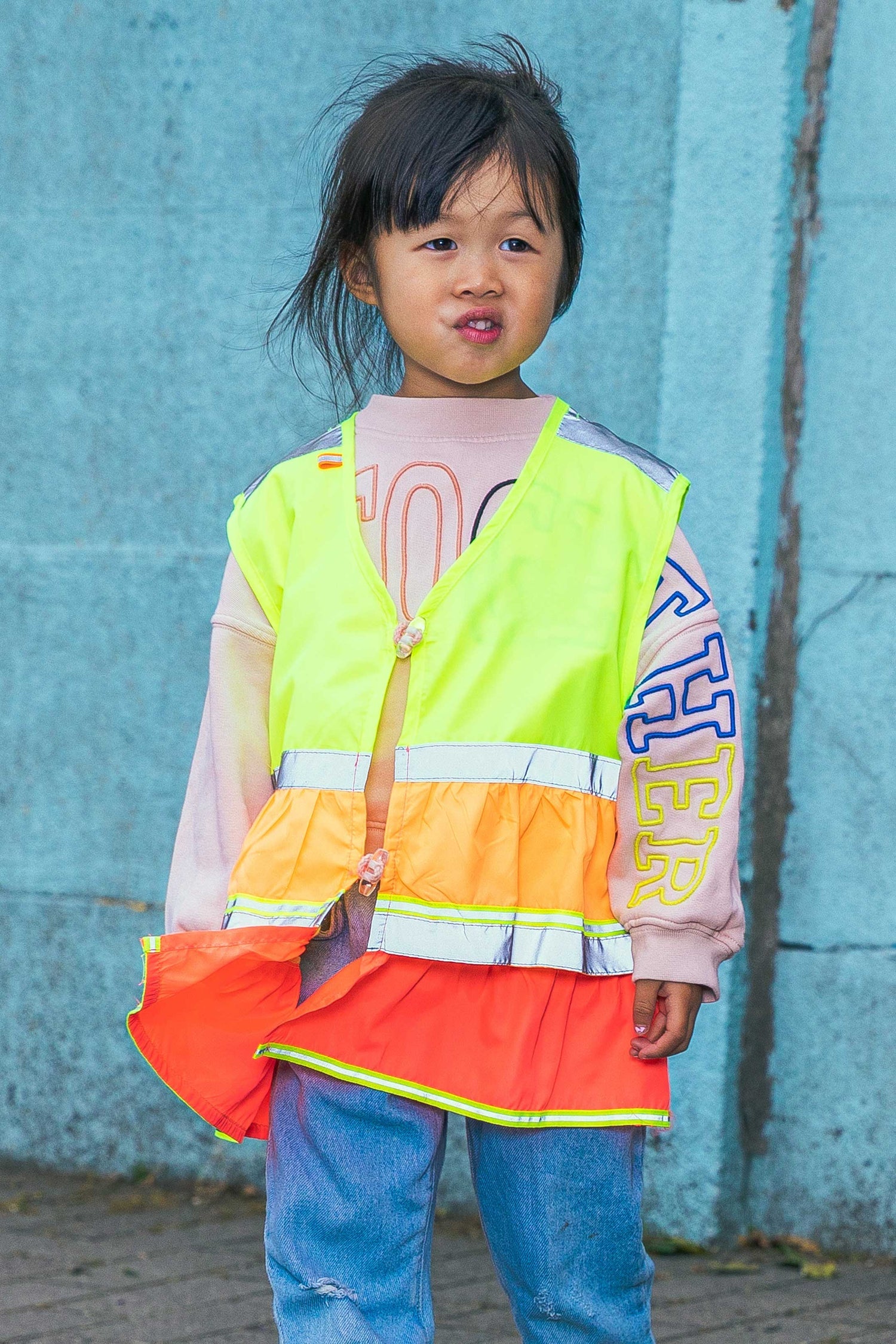 Child wearing a high-visibility safety vest over a colorful shirt against a blue wall.