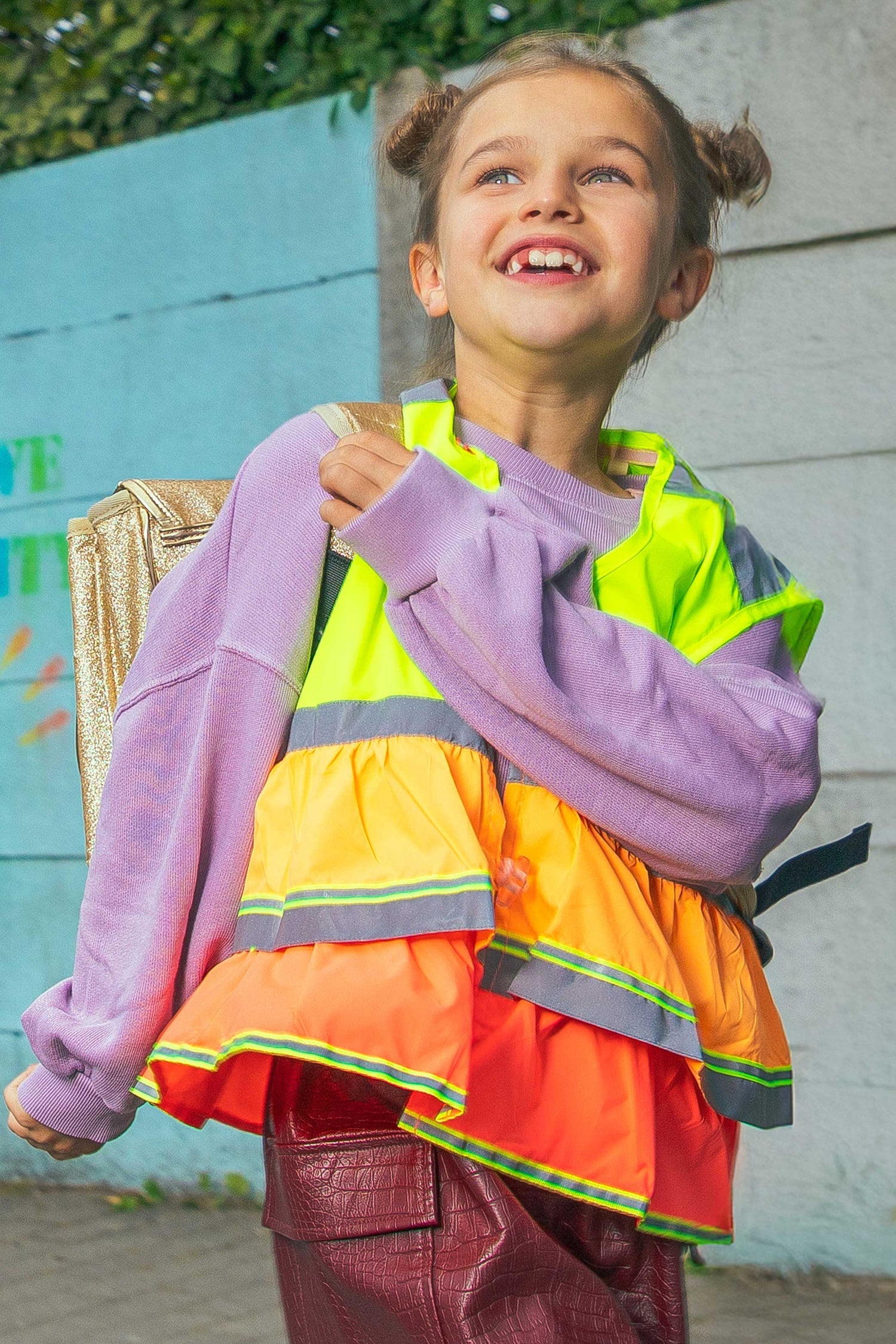 Child wearing a colorful safety vest over a layered outfit with a blue wall background