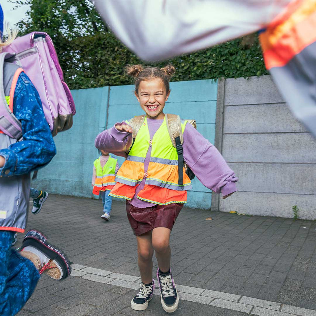 Child wearing a high-visibility vest on a sidewalk with other children in the background.