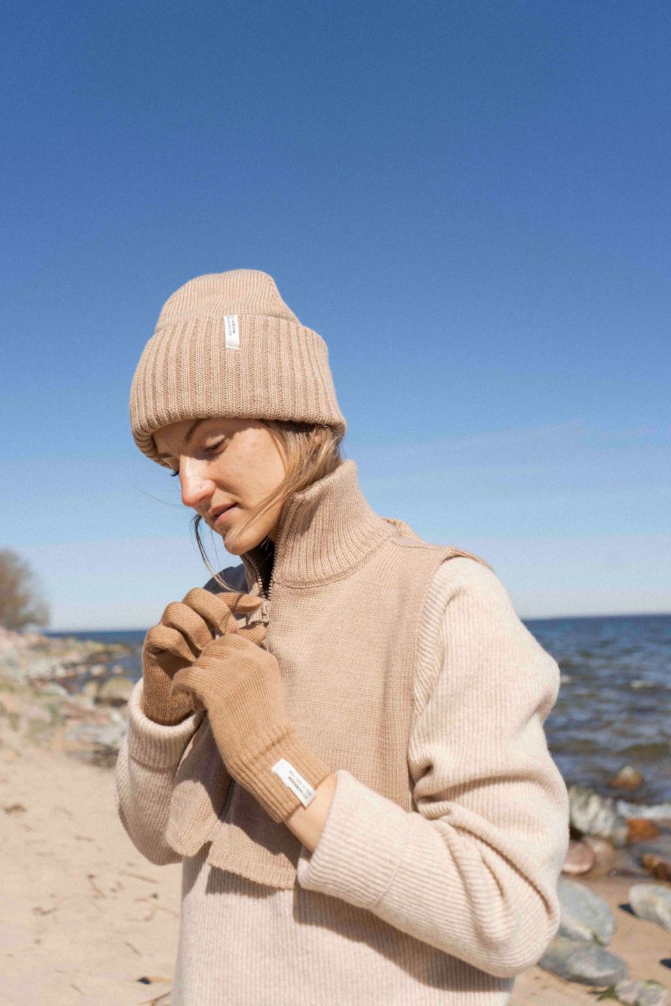 Person wearing a beige knit hat and gloves on a beach with clear blue sky