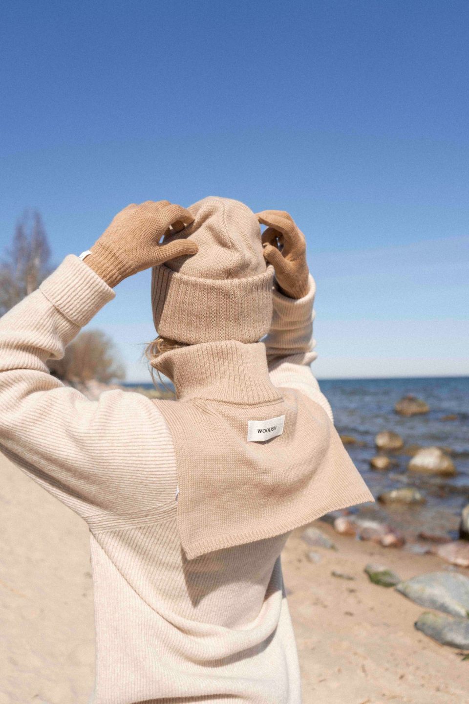 Person wearing a beige sweater and gloves adjusting a beige beanie on a beach with clear blue sky and ocean.